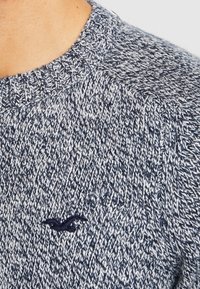 Närbild av en person som bär en texturerad marinblå och vit stickad tröja med en liten broderad fågel-logotyp på bröstet.