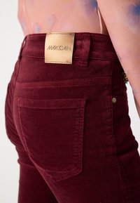 Gros plan sur une personne portant un pantalon bordeaux taille haute avec une étiquette de marque en cuir portant l’inscription « MARC CAIN » sur la ceinture.