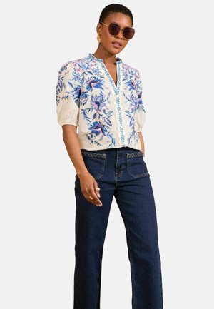 Camicia bianca con ricami floreali blu, maniche a mezza lunghezza e scollatura a V. Abbinata a jeans a campana in denim scuro, con dettagli decorativi in vita.
