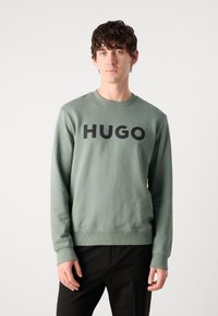 Grønn bomullssvette med rund hals, ribbestrikkede mansjetter og en stor svart "HUGO" logo over brystet, kombinert med svarte bukser.