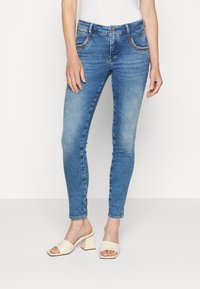 Slim-fit bleka blå jeans med rhinestone-detaljer på framfickorna, bärs med krämfärgade öppna blockklacksandaler.