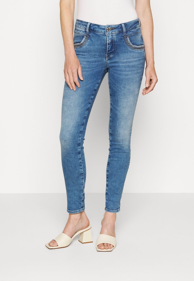 Slim-fit bleka blå jeans med rhinestone-detaljer på framfickorna, bärs med krämfärgade öppna blockklacksandaler.