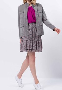 Donna vestita con giacca grigia, blusa magenta, gonna floreale e sneakers bianche, catturata a metà passo su uno sfondo uniforme.