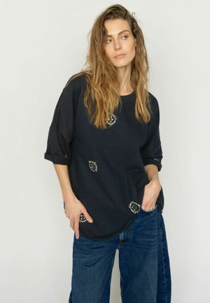 Femeie cu păr ondulat, purtând o cămașă neagră lejeră cu aplicații de strasuri și blugi albaștri, stând cu o mână în buzunar, uitându-se pe lateral.