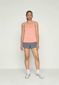  ferskenfarget tanktop med teksturert design og grå treningshorts. Modellen har på seg hvite sokker og grå joggesko med lyse detaljer.