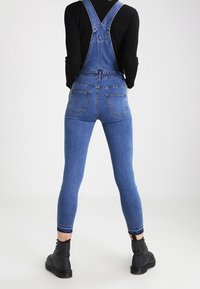 Blå denim-överaller med tajta ben, justerbara axelremmar och två bakfickor. Bärs över en svart långärmad topp och matchas med svarta stövlar.