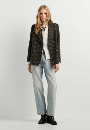 Jeune femme aux longs cheveux blonds portant un blazer sombre à fines rayures, une chemise blanche, un jean bleu clair et des bottines noires, debout sur un fond uni.
