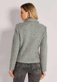 Vrouw in een lichtgrijze getailleerde blazer en donkergrijze jeans, te zien van achteren tegen een lichte, egale achtergrond.