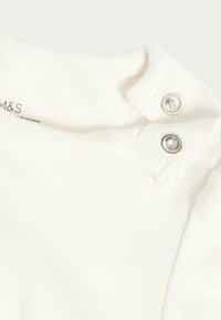 Vit ribbad baby bodysuit med tryckknappar vid axeln. Mjuk textur med ett subtilt mönster och märket synligt.