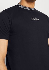 Svart bomullströja med rund halsringning, med en kontrasterande logotyp och grafik på bröstet, samt en ribbad krage med varumärke.