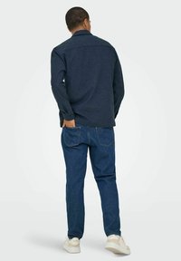 Camisa de botones azul oscuro con mangas largas, corte recto, llevada sobre jeans azul claro y zapatillas blancas; tejido suave y estilo casual.