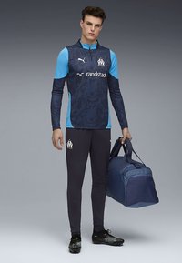 Granatowo-niebieska sportowa koszulka z długim rękawem z logo, czarne spodnie i granatowa torba sportowa, noszone z czarnymi korkami.