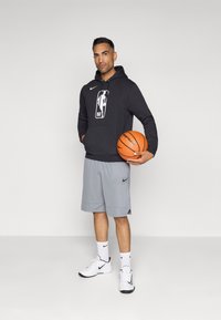 Čierna mikina s kapucňou a logom NBA, šedé nohavice, biele tenisky Nike, drží oranžový basketbal. Hladká tkanina, štýlový športový dizajn.