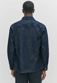 En person set fra baghovedet bærer en mørkeblå stribet skjorte, der viser teksturerede stofdetaljer mod en ensfarvet lys baggrund.