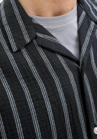 Czarna i szara koszula w paski z teksturowanej tkaniny, z wyciętym kołnierzem i detalami z guzikami. Pod spodem widoczny szary t-shirt z okrągłym dekoltem.