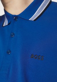 Blå polo-skjorta med en texturerad tyg, med en kontrasterande vit och svart randig krage och en svart broderad logotyp. Fyra knappar synliga.