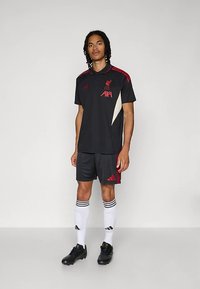 Man som står i svart Liverpool fotbollskit med röda detaljer, vita knästrumpor och svarta fotbollsskor mot en enkel grå bakgrund.