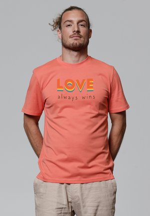 Koraalkleurig t-shirt met korte mouwen, voorzien van meerkleurige tekst "LIEFDE wint altijd" gedrukt in het midden. Zacht, katoenen materiaal.