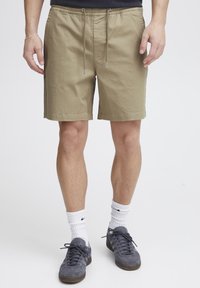 Shorts beiges en coton avec taille élastique et cordon de serrage. Ils disposent de poches latérales et d'une coupe décontractée, portés avec des baskets grises et des chaussettes blanches.