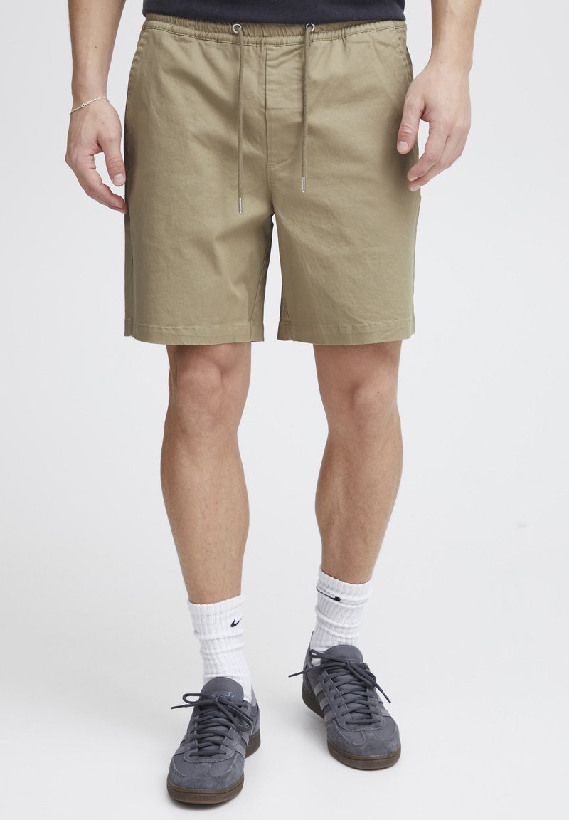 Shorts beiges en coton avec taille élastique et cordon de serrage. Ils disposent de poches latérales et d'une coupe décontractée, portés avec des baskets grises et des chaussettes blanches.