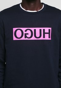 Felpa blu navy con un grande logo rettangolare rosa con la scritta "HUGO" in nero. Collo a giro e morbidezza al tatto.