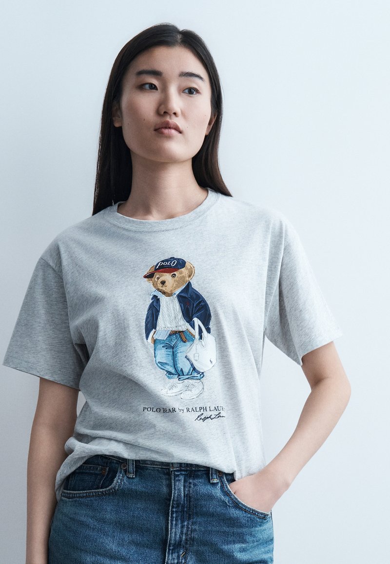 Νεαρή γυναίκα που φοράει ένα ανοιχτό γκρι t-shirt με τυπωμένη μια αρκουδάκι ντυμένο με casual ρούχα και μπλε τζιν, στέκεται με το χέρι στην τσέπη.