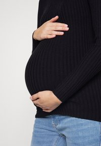 Personne enceinte portant un pull noir côtelé et un jean bleu, caressant doucement son ventre avec les deux mains.