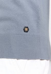 Lichtblauwe gebreide sweater met geribbelde zoom en een klein hexagonaal logo met drie horizontale lijnen in goud en zwart.