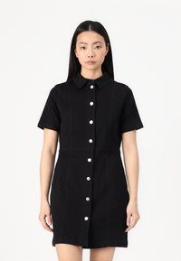 Vestido preto de manga curta feito de denim, com fecho frente em botão, colarinho e bolsos nas costuras laterais. Apresenta botões prateados e uma silhueta ajustada.