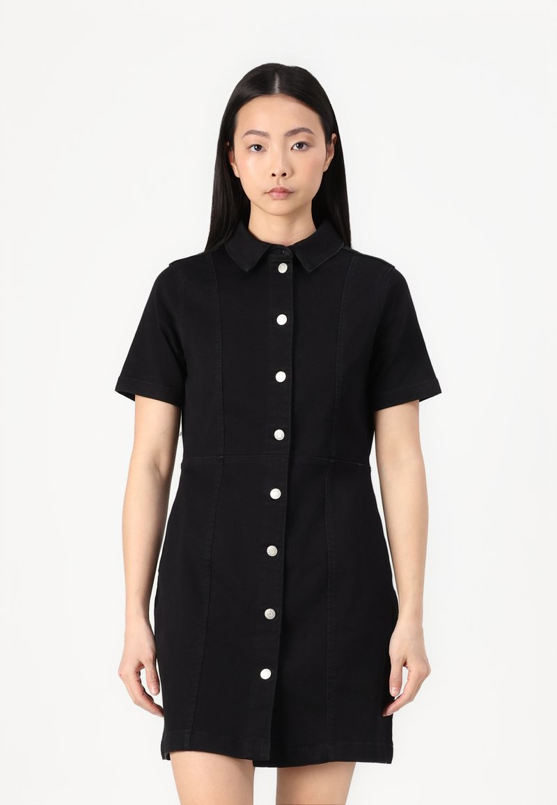 Vestido preto de manga curta feito de denim, com fecho frente em botão, colarinho e bolsos nas costuras laterais. Apresenta botões prateados e uma silhueta ajustada.