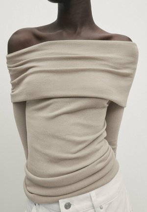 Personne à la peau foncée portant un pull beige côtelé à col bateau et un pantalon blanc sur un fond uni.