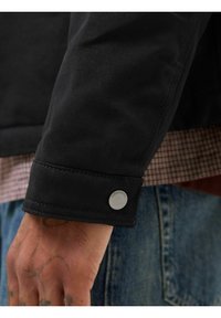 Černý plátěný rukáv bundy s detaily na patentkách. Viditelná kostkovaná košile pod ní a džíny v pase. Detail texturované látky.