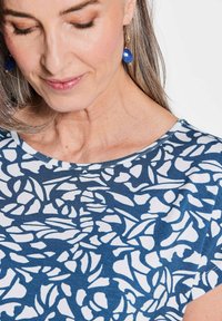 Blau-weißer Oberteil mit Blumenmuster, kurzärmelig, mit runder Ausschnitt und glatter Textur, getragen mit herabhängenden blauen Ohrringen.