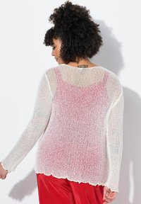 Maglione lavorato a maglia bianco con un orlo a scallop, caratterizzato da una texture trasparente e maniche lunghe, indossato sopra un top rosa e abbinato a pantaloni rossi.