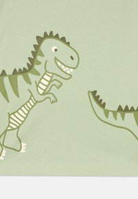 Ανοιχτό πράσινο T-shirt με παιχνιδιάρικα γραφικά δεινοσαύρων σε διάφορες αποχρώσεις του πράσινου, με ξεχωριστά δόντια και αγκάθια, σε μαλ ακό ύφασμα.