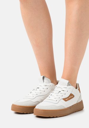 Witte lage sneakers met bruine rubberen zolen, gedragen op blote benen, met "Marc O'Polo" branding op het zijpaneel.