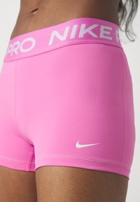 Lyserøde sports-shorts med en bred talje, som har "NIKE PRO" i hvid. Stoffet er glat, og der er et lille hvidt Nike-logo på siden.
