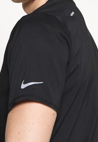 Černé tričko s krátkým rukávem z texturované látky, s reflexním logem Nike na levém rukávu a minimalistickým designem.