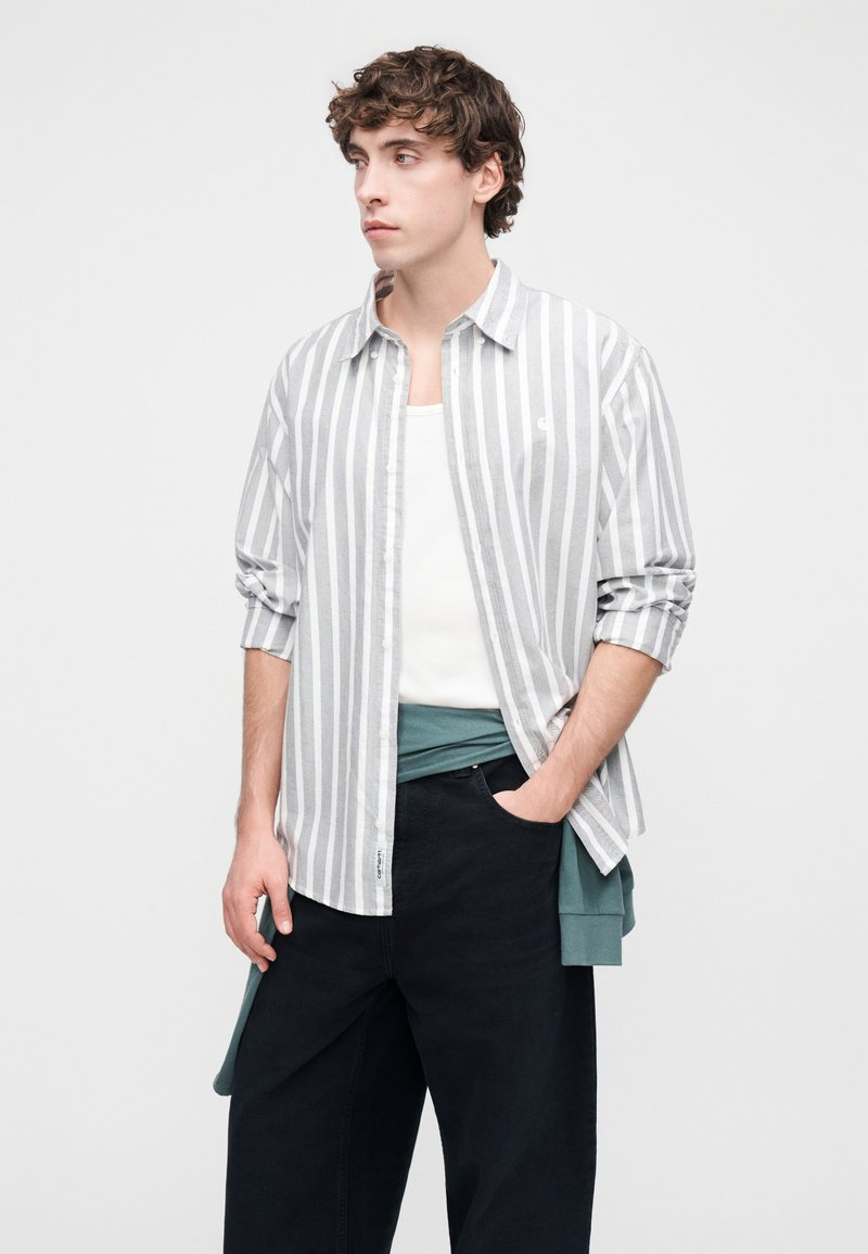 Camicia da uomo leggera a righe grigie e bianche con bottoni, maniche lunghe, indossata sopra una maglia turchese, abbinata a pantaloni scuri.