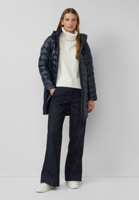 Puhasto plašč v temno modri barvi, z kapuco. Bel teksturiran pulover, visokega ovratnika. Razširjene temne kavbojke. Bele superge z debelim podplatom. Enobarvna ozadje.