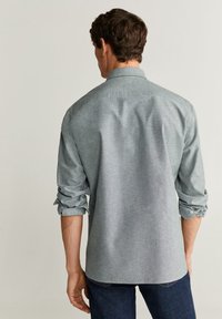 Camisa de botones gris claro con mangas enrolladas, hecha de una tela suave, con cuello estándar y dobladillo recto, llevada sobre jeans oscuros.