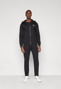 Μαύρο φούτερ με φερμουάρ μπροστά και τσέπη, με λογότυπο, συνδυασμένο με εφαρμοστά φούτερ παντελόνια, φορεμένο με λευκά αθλητικά παπούτσια.