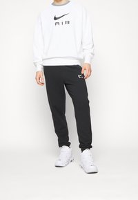 Vit sweatshirt med svart "AIR"-text och grå krage; svarta joggers med vitt Nike-logotyp. Båda är gjorda av mjukt material och bärs med vita sneakers.