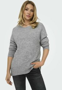 Suéter de punto gris con un dobladillo acanalado, cuello en V y puños enrollados. Textura suave y corte relajado, combinado con jeans oscuros.