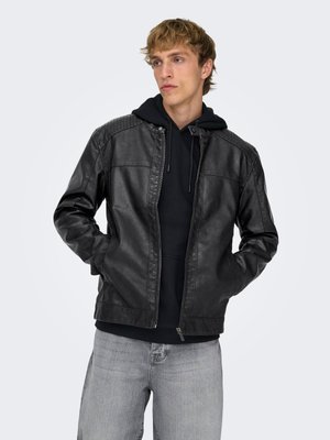 Jovem usando um casaco de couro preto por cima de um hoodie preto e jeans cinza claro, em pé com as mãos nos bolsos do casaco.