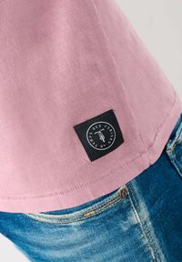 T-shirt in cotone rosa chiaro con un'etichetta nera che presenta testo bianco. La maglietta ha un orlo arrotondato ed è abbinata a jeans di denim blu sbiaditi.