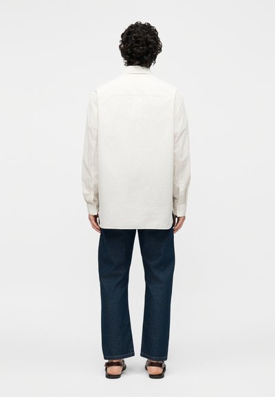 Άνδρας με κατσαρά μαλλιά που φοράει μακρυμάνικο κρεμ πουκάμισο, σκούρα μπλε τζιν και καφέ σανδάλια, στέκεται με την πλάτη προς τα εμπρός σε μονόχρωμο φόντο.