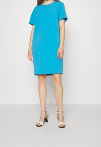 Tyrkysové šaty s krátkým rukávem, kulatým výstřihem, rovného střihu a jemné textury. Nošeny s podpatky s kotníkovým páskem v bílé a černé barvě.