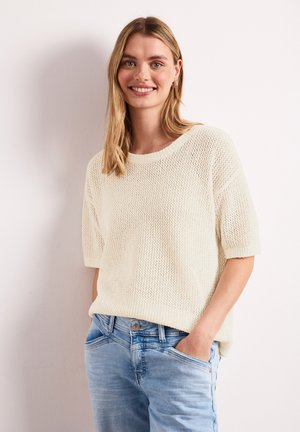 Femme souriante aux cheveux blonds porte un pull blanc tricoté ample à manches courtes et un jean bleu clair, debout, une main dans la poche.