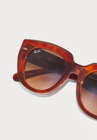 Gafas de sol de ojo de gato con un marco ámbar translúcido, lentes marrones degradados y un acento de logo plateado. Textura suave y forma audaz.